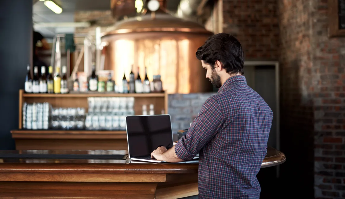 Brewery owner reviewing marketing performance on a laptop at the taproom bar during the day