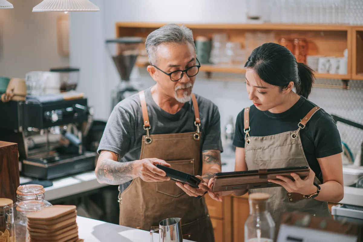 A café owner reviewing marketing performance on a tablet at the counter while a barista serves customers