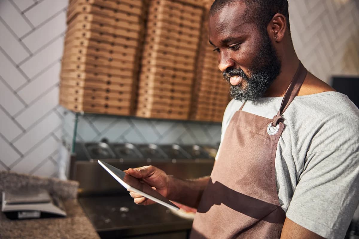 Pizzeria owner reviewing online orders on a tablet at the counter of a busy pizza shop