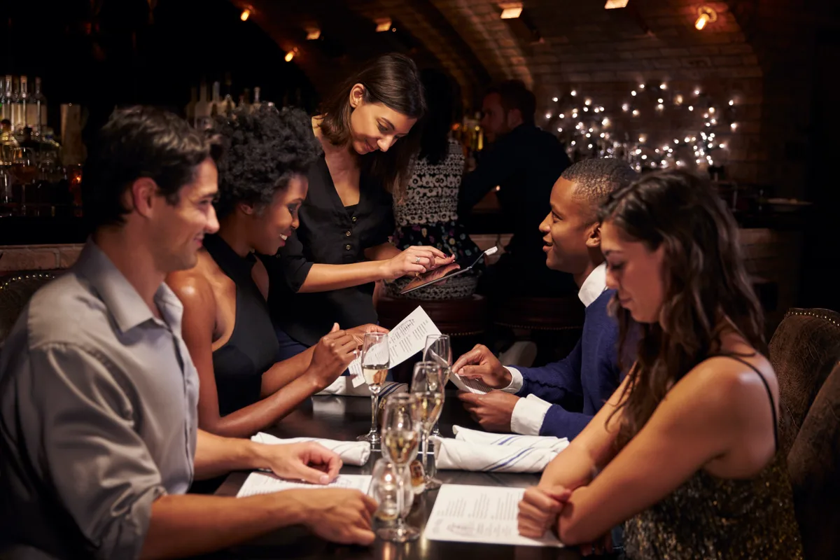 A full restaurant dining room during dinner service with a server taking an order at a table