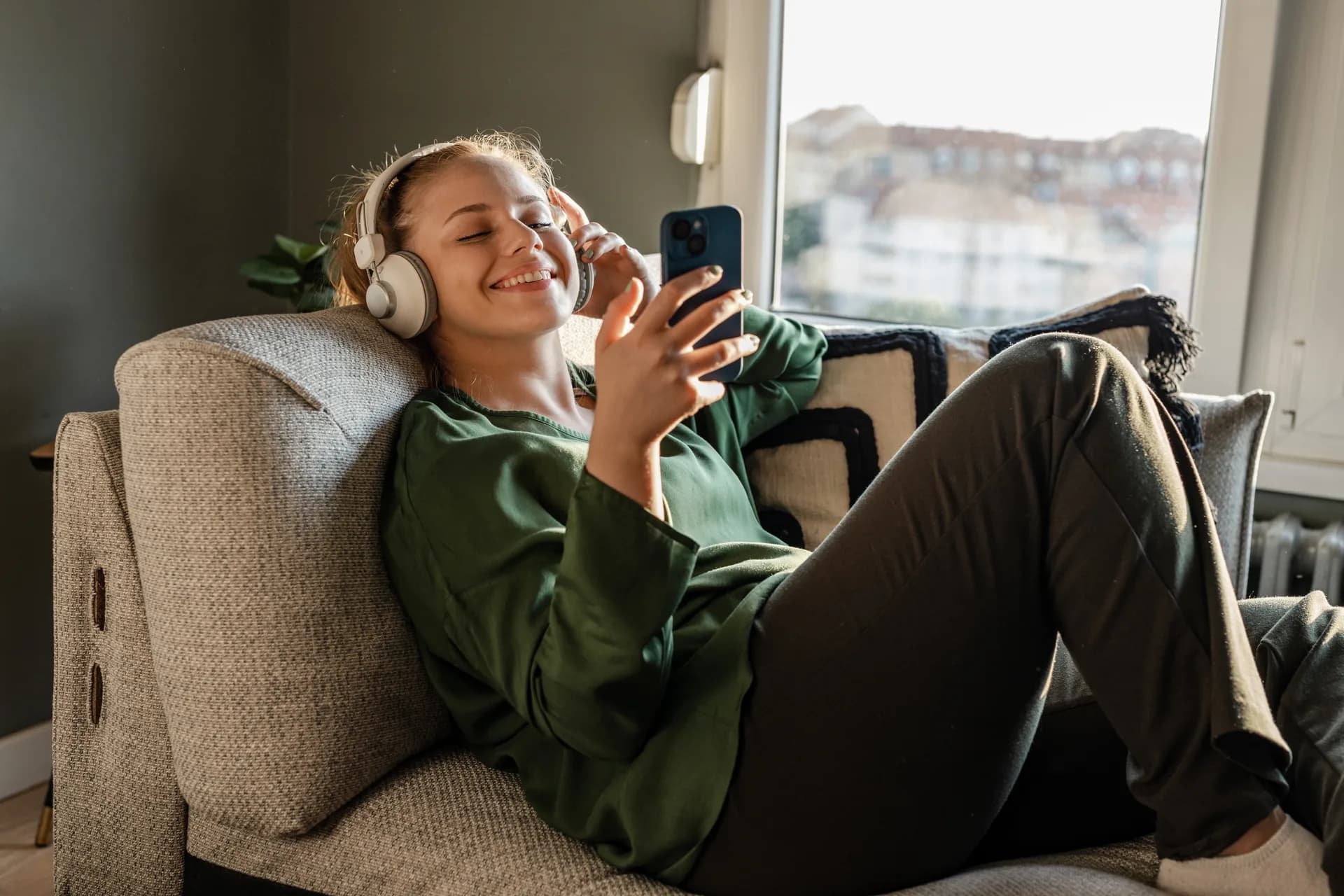 A young woman with white headphones, smiling at her phone in a sunlit room