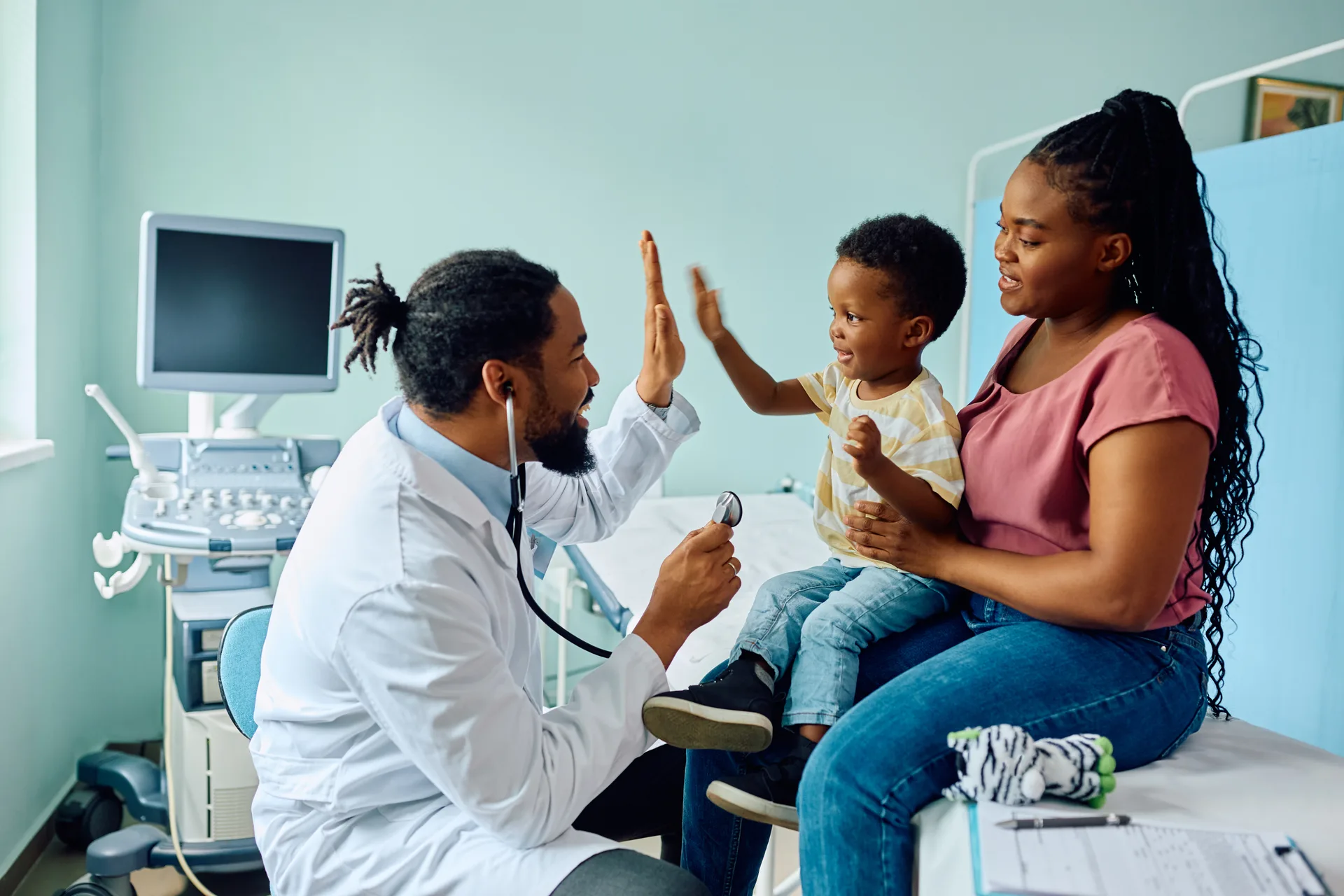 A pediatrician giving a young patient a high-five during a clinic visit, mom watching on