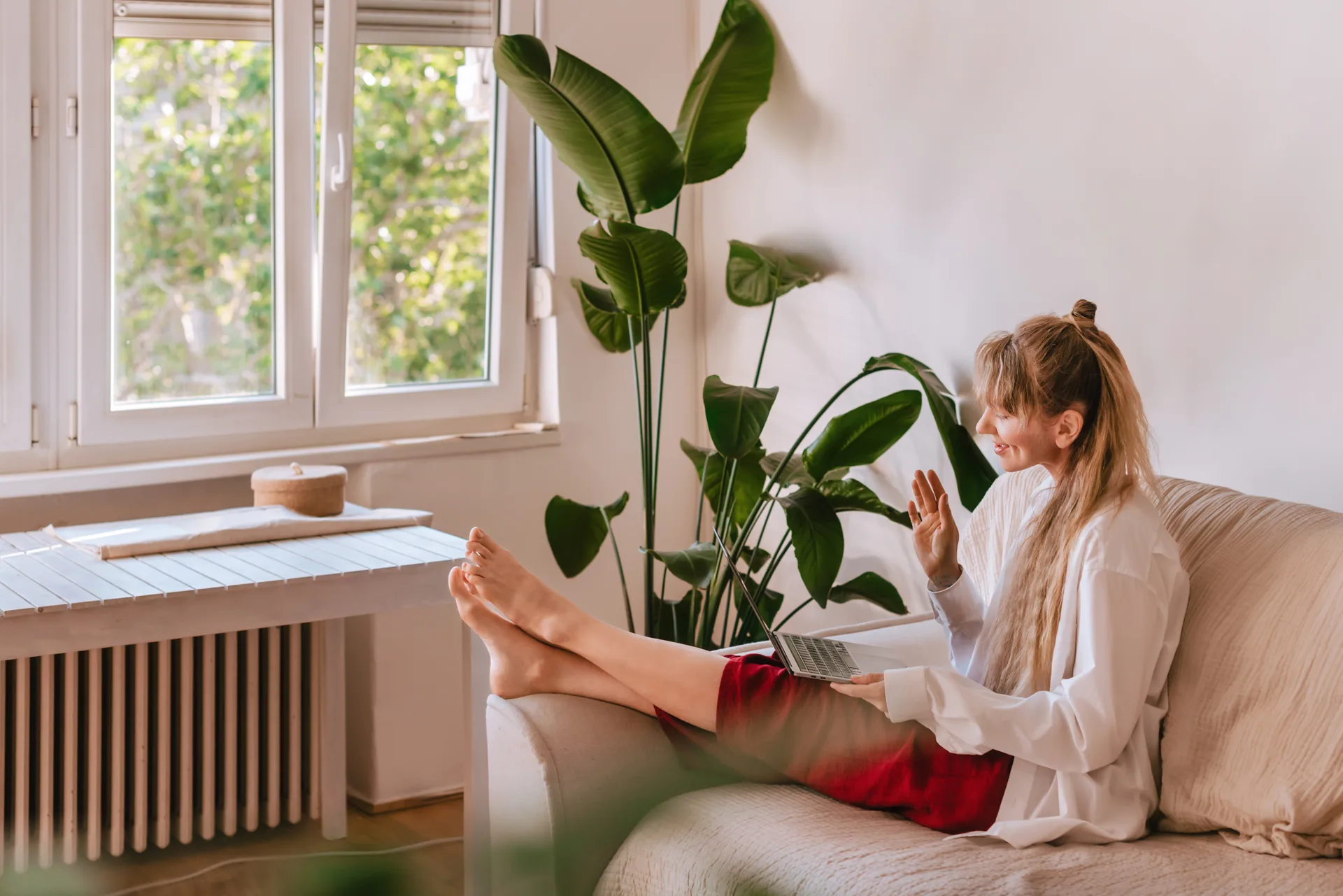 A woman on a bright sofa waving hello to her therapist on a laptop