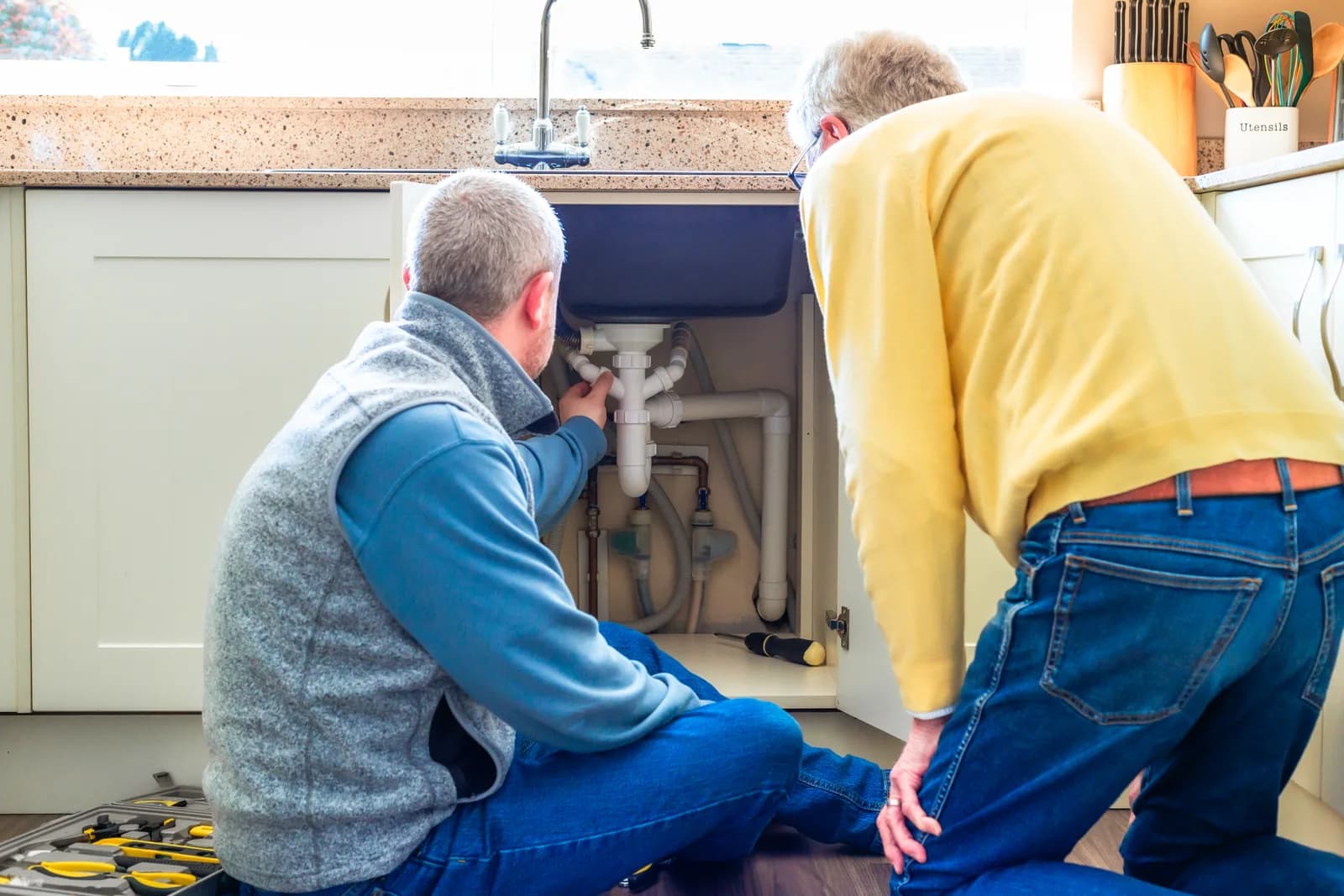 Plumber repairing pipes under a kitchen sink while a homeowner looks on